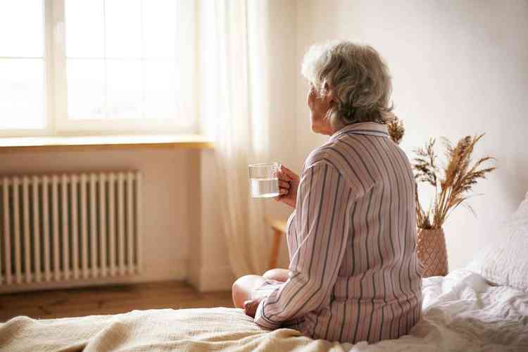 Senhora da terceira idade sentada em sua cama segurando uma xcara de ch