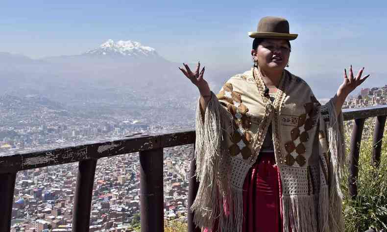 com chapu, saia e xale tpicos bolivianos, Alwa ergue as mos, com os Andes ao fundo