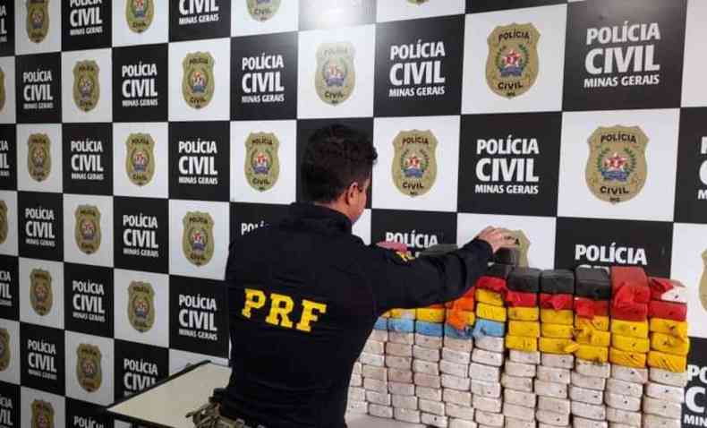 Policial com uniforma da PRF empilha barras de pasta base de cocaína; parede ao fundo mostra logomarca e brasão da Polícia Civil