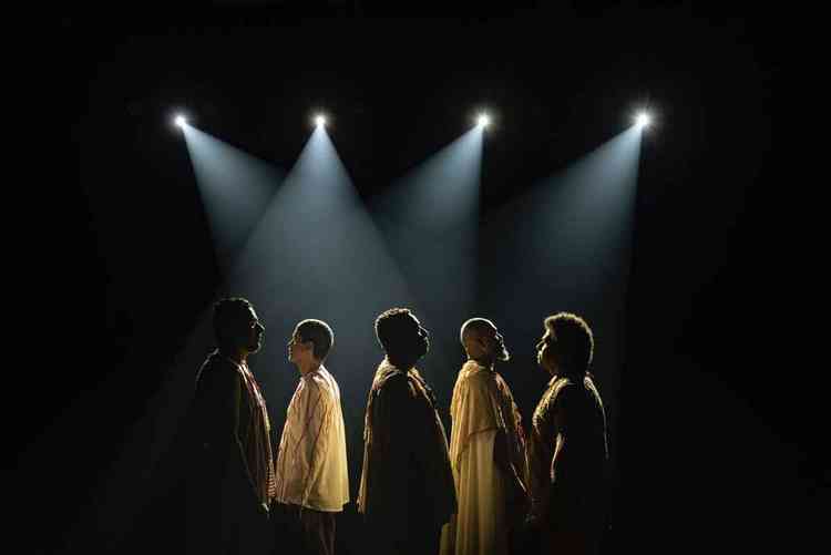 Os integrantes do Cordel do Fogo Encantado, de p, no palco, iluminados por holofotes
