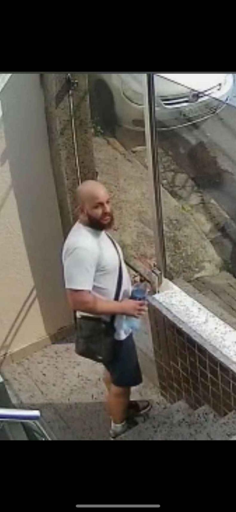 Homem careca de blusa branca, barba e bolsa preta, saindo de portaria de apartamento 