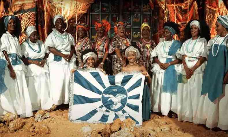 Mulheres negras com vestidos brancos em torno da bandeira da Portela 