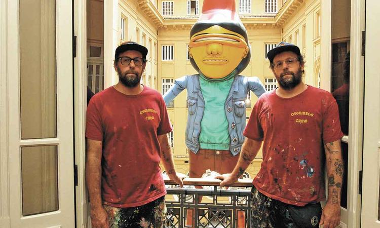 Os Gmeos Gustavo e Otvio Pandolfo olham para a cmera. Atrs est um boneco criado por eles
