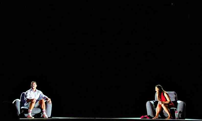 Os atores Priscila Fantin e Bruno Lopes sentados em poltronas da cor cinza, cada um em um canto do palco, riem, em cena de 'Precisamos falar de amor sem dizer eu te amo' 
