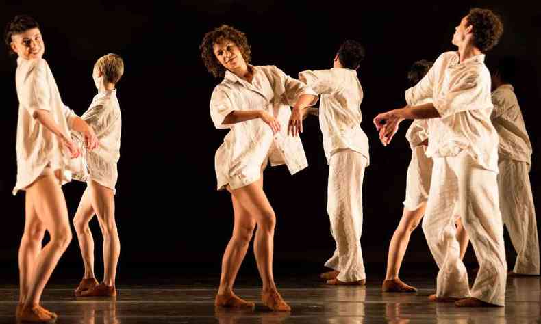 Vestindo roupas brancas, sete bailarinos do Grupo Corpo danam no palco. Duas bailarinas sorriem para a cmera