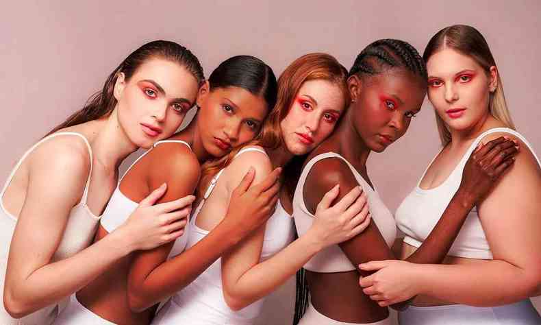 Cinco mulheres de diferentes tons de pele usando camisetas brancas apoiando umas nas outras 