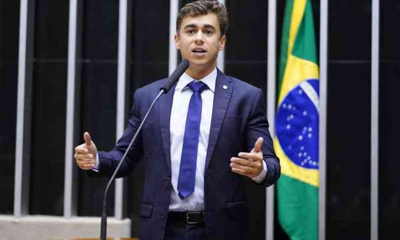 Nikolas Ferreira na Plenria da Cmara dos Deputados falando ao microfone. Ao fundo uma bandeira do Brasil. Ele  um jovem branco de cabelos lisos e usa terno
