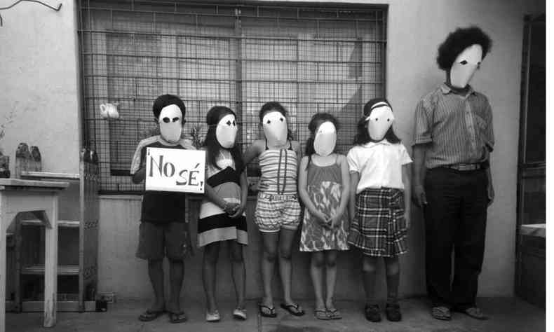 Cinco crianças e um adulto usando máscaras estão enfileiradas, à frente de janela com grades. O primeiro menino da fila segura cartaz onde se lê No sé, que significa não sei