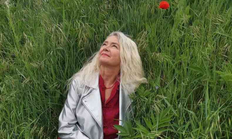 de vestido vermelho e jaqueta prateada, sentada em meio à vegetação, Virginie olha para cima 