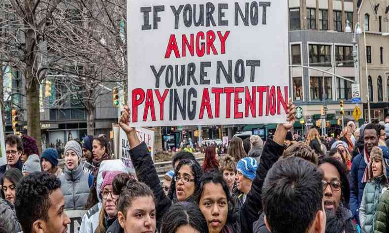 multido em protesto, onde uma mulher segura um cartaz escrito: 'If you're not angry, you're not paying attention'.