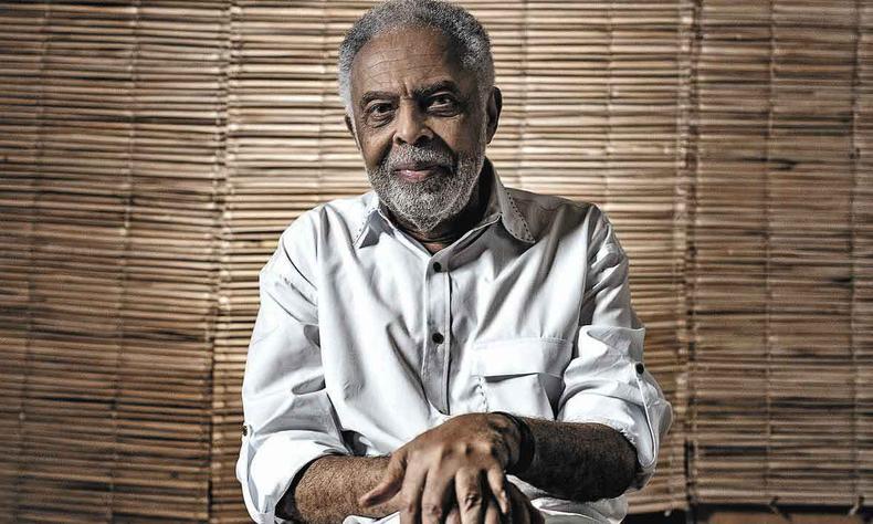 Sentado e com as mos cruzadas, Gilberto Gil sorri e olha para a cmera