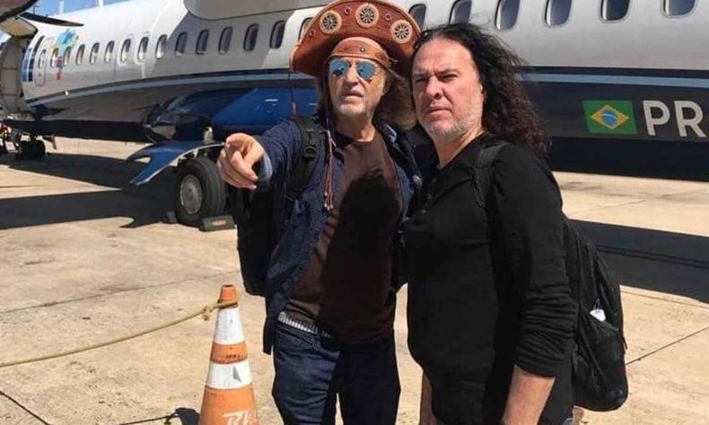 Alceu Valena, usando chapu de vaqueiro, aponta para a frente, ao lado do guitarrista Paulo Rafael, na pista de um aeroporto. Ao fundo, v-se um avio