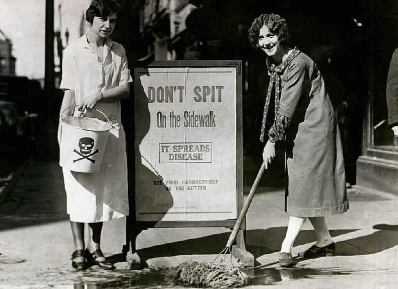 Em foto em preto e branco, duas mulheres ao lado de um cartaz onde se l:''no cuspa na calada, isso espalha doenas'