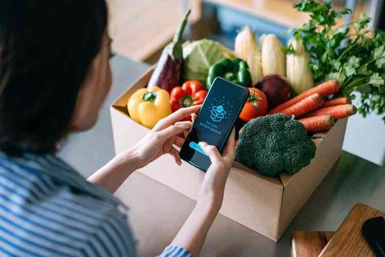 Mulher de costas mexendo no celular perto de caixa com frutas, legumes e verduras