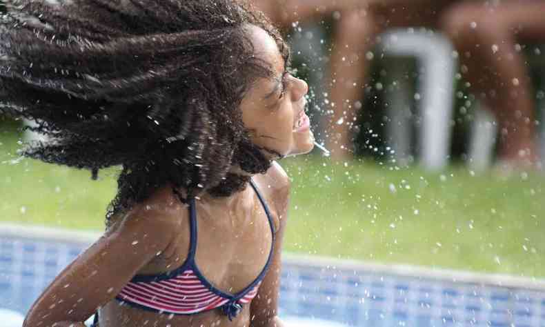 Uma menina negra, saindo da piscina e balançando seus cabelos enquanto sorri