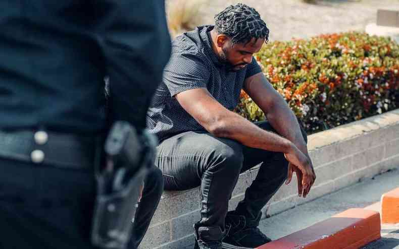 Homem negro sentado no meio-fio observado por um policial