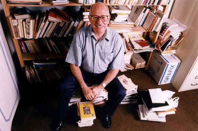 O escritor Silviano Santiago sorri para a cmera, sentado numa pilha de livros,  frente de uma estante toda bagunada