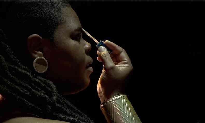 De perfil e usando pulseira de motivos africanos, a compositora Sheruba Déluz pinta a sobrancelha 