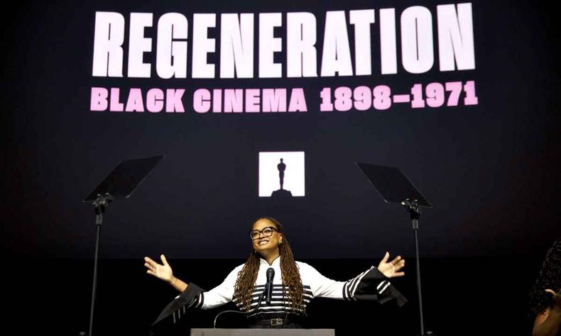 Cineasta negra Ava DuVernay abre os braos em frente a pequeno parlatrio. Ao fundo v-se a imagem do Oscar e cartaz da exposio Regenerao: o cinema afro-americano 1898-1971