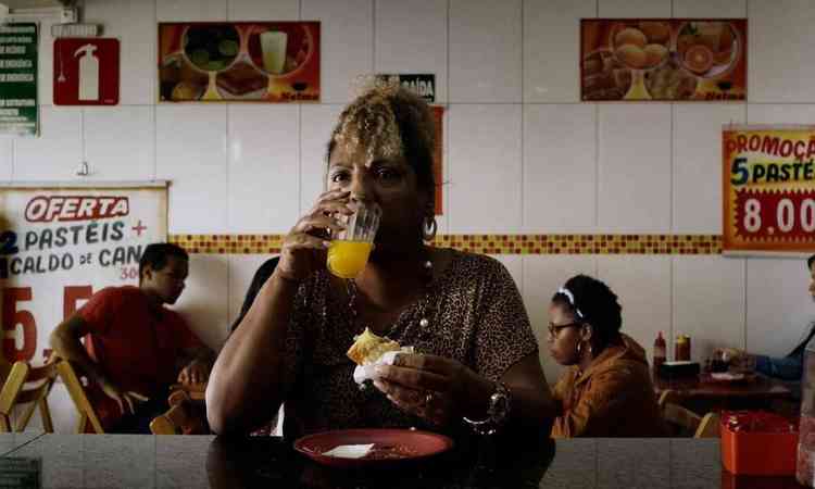 Atriz Rejane Faria come pastel e bebe laranjada, sentada  mesa de restaurante popular, em cena do filme Marte Um