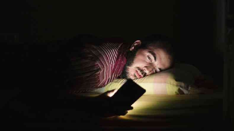 Homem branco de cabelo preto na cama, com um fundo escuro, olha para a tela de um celular