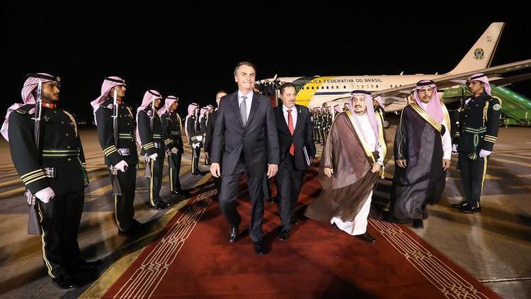 Bolsonaro saindo de avião brasileiro em tapete vermelho, rodeado por homens com trajes típicos da Arábia Saudita