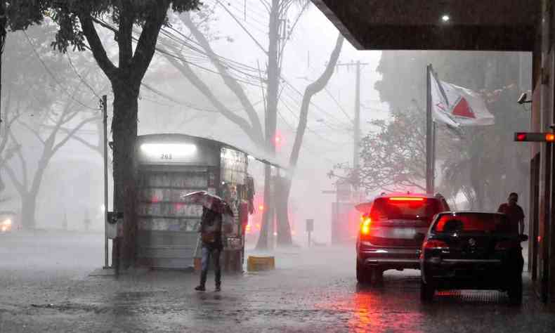 Pessoa na chuva com um guarda-chuva prximo a uma banca de jornal, carros e uma bandeira do estado de Minas Gerais