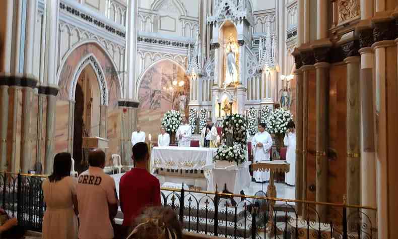 Foto dentro da igreja, da para ver as pessoas assistindo as missas, com Nossa Senhora ao fundo. A cor branca dita a cor do fundo junto ao mrmore bege