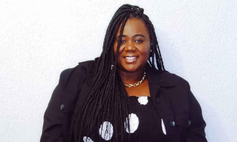 Jane Luciana, mulher negra usando vestido preto com crculos brancos e um blazer preto por cima, sorri para a cmera 