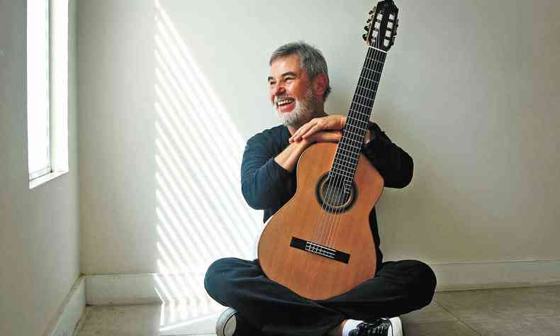 Sentado no cho, violonista Marco Pereira sorri, tendo ao fundo a luz do sol que atravessa uma janela 
