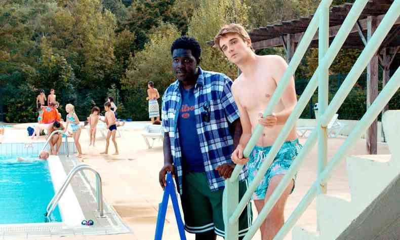 Um jovem negro e um jovem branco olham para o lado, tendo ao fundo pessoas à beira da piscina, no filme 'Embarque