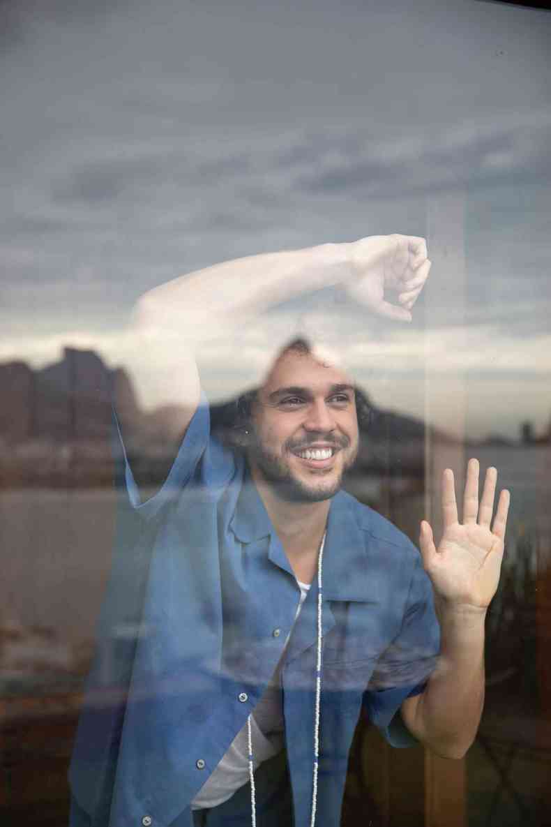 Theo Bial encosta a testa no próprio braço, apoiado a uma parede de vidro, e sorri