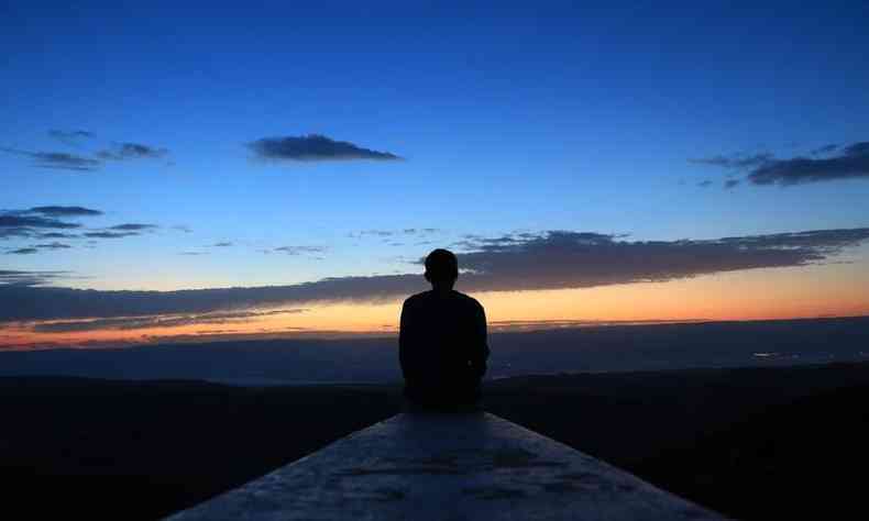 Homem sentado de costas, numa passarela, olhando o horizonte em um entardecer