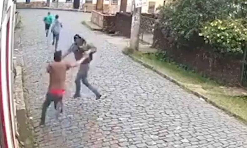Homem de verde e cinza correndo enquanto um de preto e outro de marrom brigam na rua de pedra