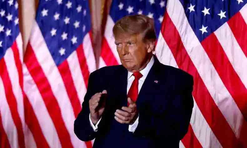 Na foto, Donald Trump, ex-presidente dos Estados Unidos; no momento, ele aplaude em frente a bandeiras dos EUA, usa um terno preto e gravata vermelha