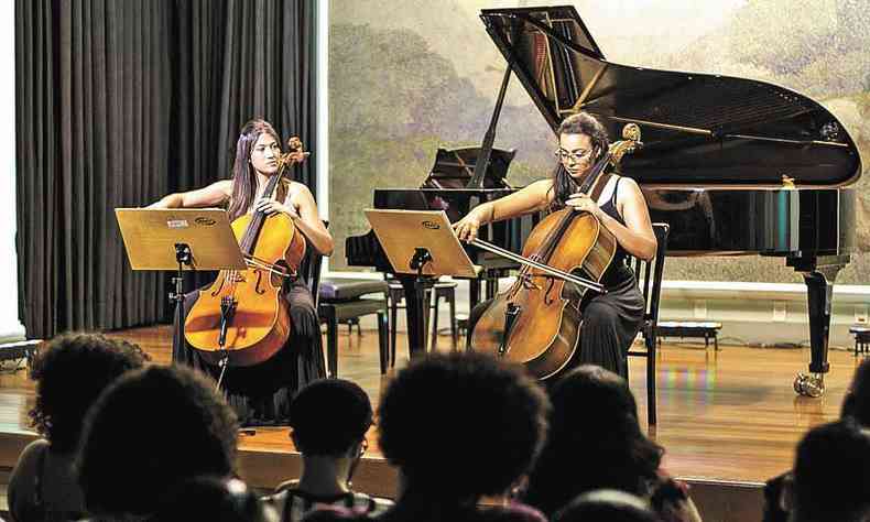 As violoncelistas Ana Paula Rocha e Talitha Marinho tocam seu instrumento numa sala, cercadas por plateia