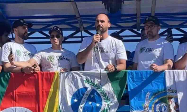 Eduardo bolsonaro falando em microfone em evento usando uma camisa branca onde se l 'pro armas'