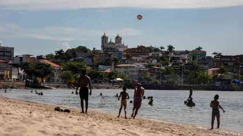 Pessoas em praia de Salvador jogando altinha, com igreja e casas ao fundo