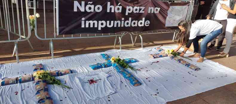 Cruzes formadas por sacos de feijo no cho, mulher no canto direito deixa flores no local. Gradis ao fundo com uma faixa escrito 'No h paz na impunidade'