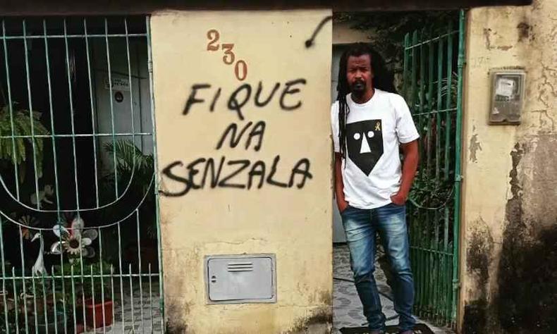 Muro da casa do candidato a deputado federal Mazo (PSB) foi pichado na madrugada de terça-feira (20/9)(foto: Instagram @mazo4041/Reprodução) Foto do muro pichado da casa de Mazo. A parede é amarela e lê-se nela 'Fique na senzala' abaixo do número da casa, 230. Ao lado do muro e em frente ao portão, está Mazo, um homem negro com dreadlocks longos nos cabelos escuros. Ele usa uma camiseta branca com a estampa do logo da banda Baiana System, que é uma máscara 2D com formas geométricas.