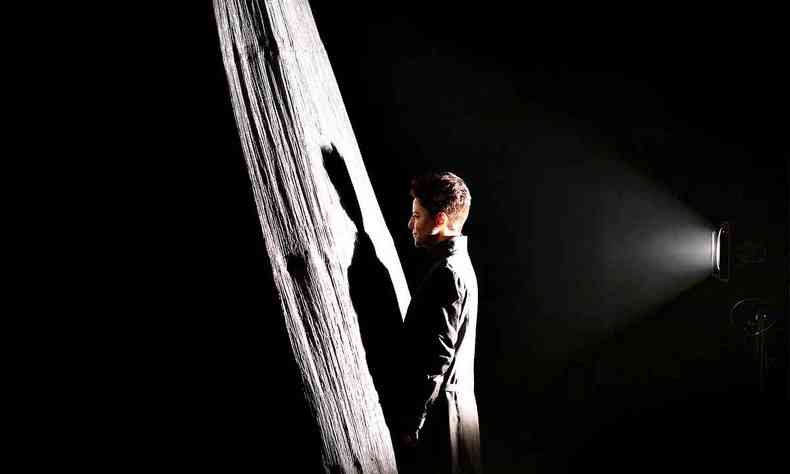 Fernanda Takai, de p, vestida com casaco cinza, tem sua sombra projetada pela luz de um refletor em uma estrutura vertical  sua frente