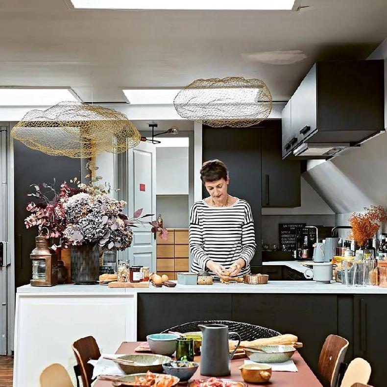 Fotografia colorida mostra uma bancada de uma cozinha aberta para a sala e uma mulher branca cozinhando