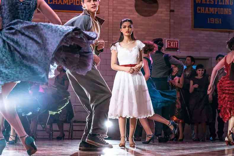 De vestido branco, a atriz Rachel Zegler, com o rosto triste, está no meio de dançarinos num salão de baile, no filme Amor, sublime amor