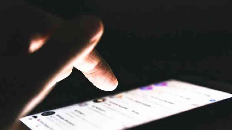 Fotografia colorida mostra uma mo tocando em uma tela de celular com um fundo escuro