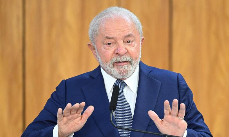 Lula com as mos espalmadas a sua frente. usa Terno azul, camisa branca e gravata tambm azul. A parede atrs de Lula de madeira clara 