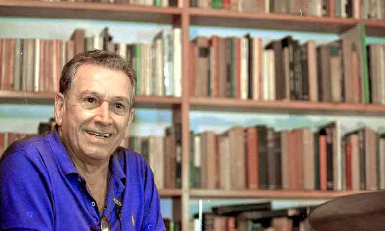 Fernando Sabino sorri e ao fundo v-se estante com prateleiras repletas de livros