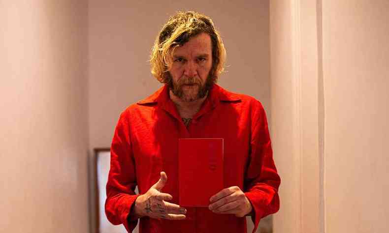 Com vestido vermelho, de  num corredor, Otto segura o seu livro vermelho 