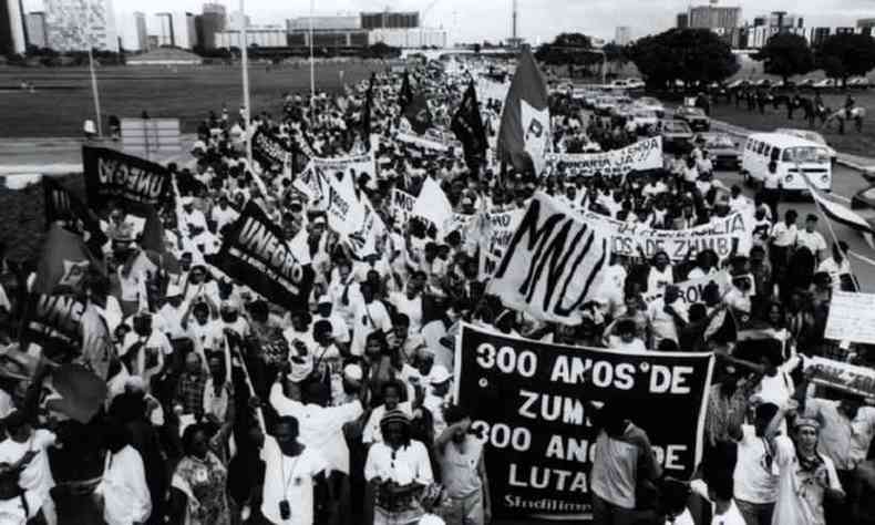 Foto em preto e branco de vrias pessoas, maioria negra, em uma marcha em prol de polticas pblicas para a populao negra em novembro de 1995. As pessoas seguram cartazes e se organizam em frente a um gramado