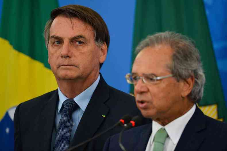 Paulo Guedes ao lado de Bolsonaro com a bandeira do Brasil ao fundo 