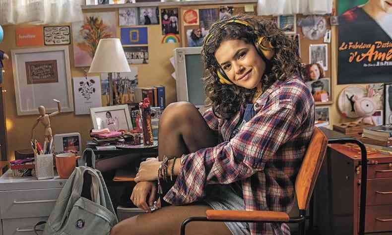 De camisa xadrez, fones de ouvido, sentada numa cadeira, Maisa Silva grava cena de 'de volta aos 15'